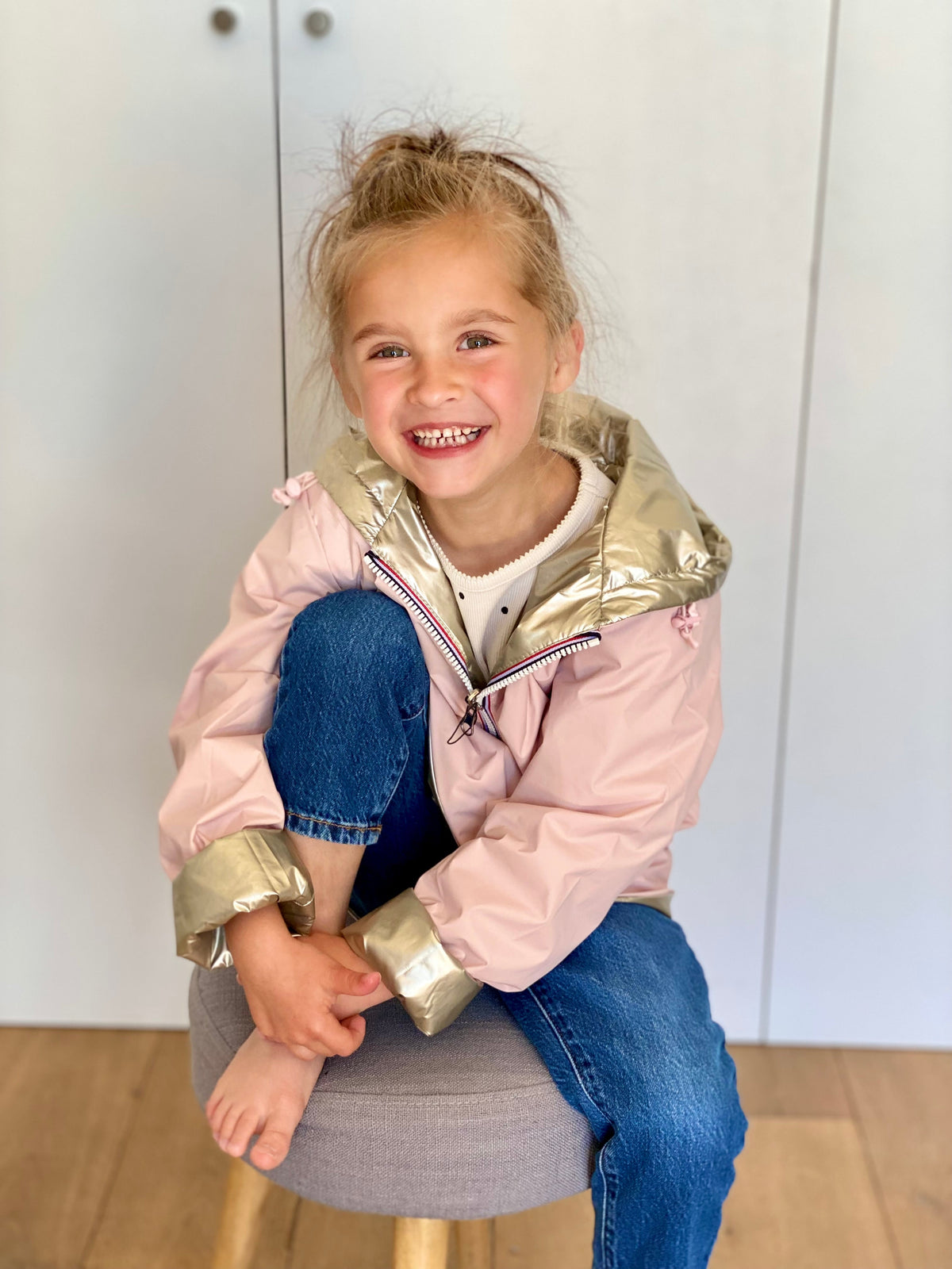 Une fille souriante en imperméable rose et doré assise sur un pouf, tenant son pied posé sur le tabouret, avec un jean bleu visible à côté.