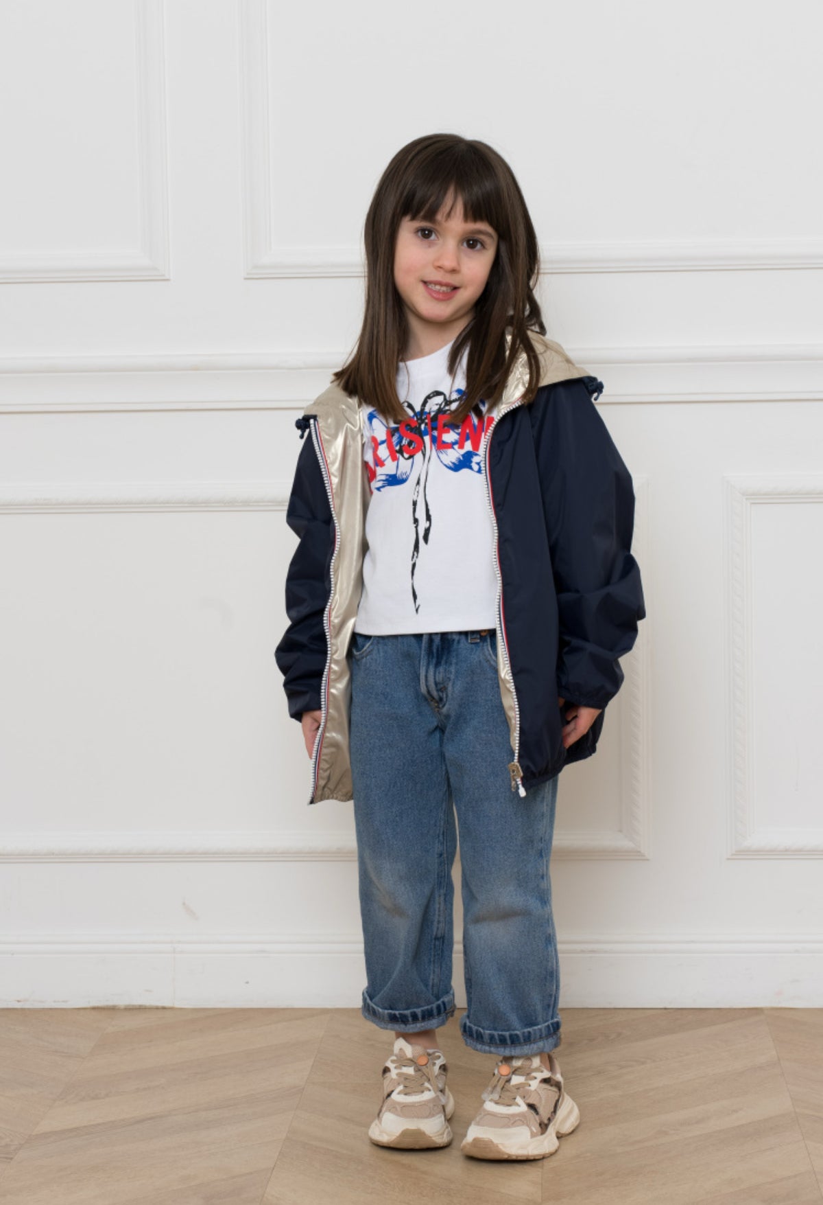 Jeune fille portant l'imperméable réversible léger GARANCE KIDS de LAURA JO, avec capuche et poches zippées, debout devant un mur blanc.