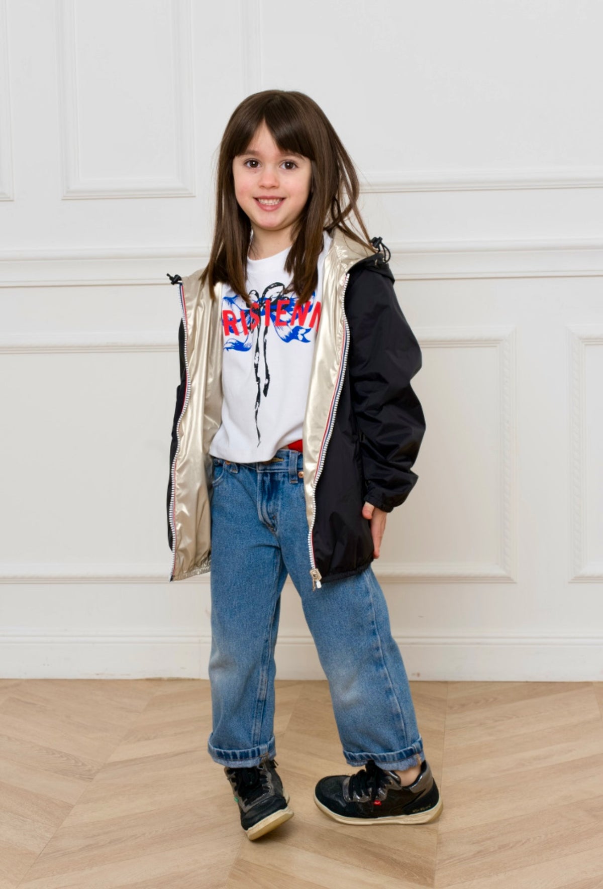 Une fille porte l'imperméable réversible GARANCE KIDS LAURA JO avec capuche et poches, debout devant un mur blanc.