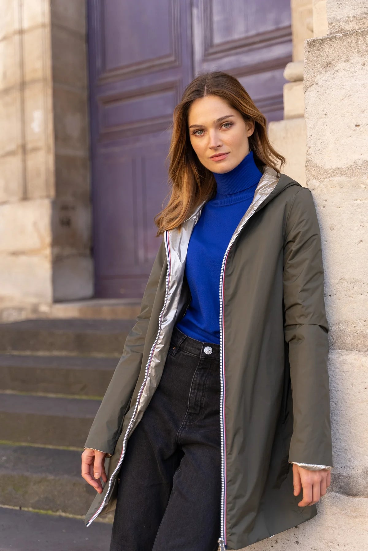 Femme portant l'Imperméable réversible rembourré LOUISON LONG - Kaki/Doré de Laura Jo, appuyée contre un mur, montrant la texture et le style du manteau.