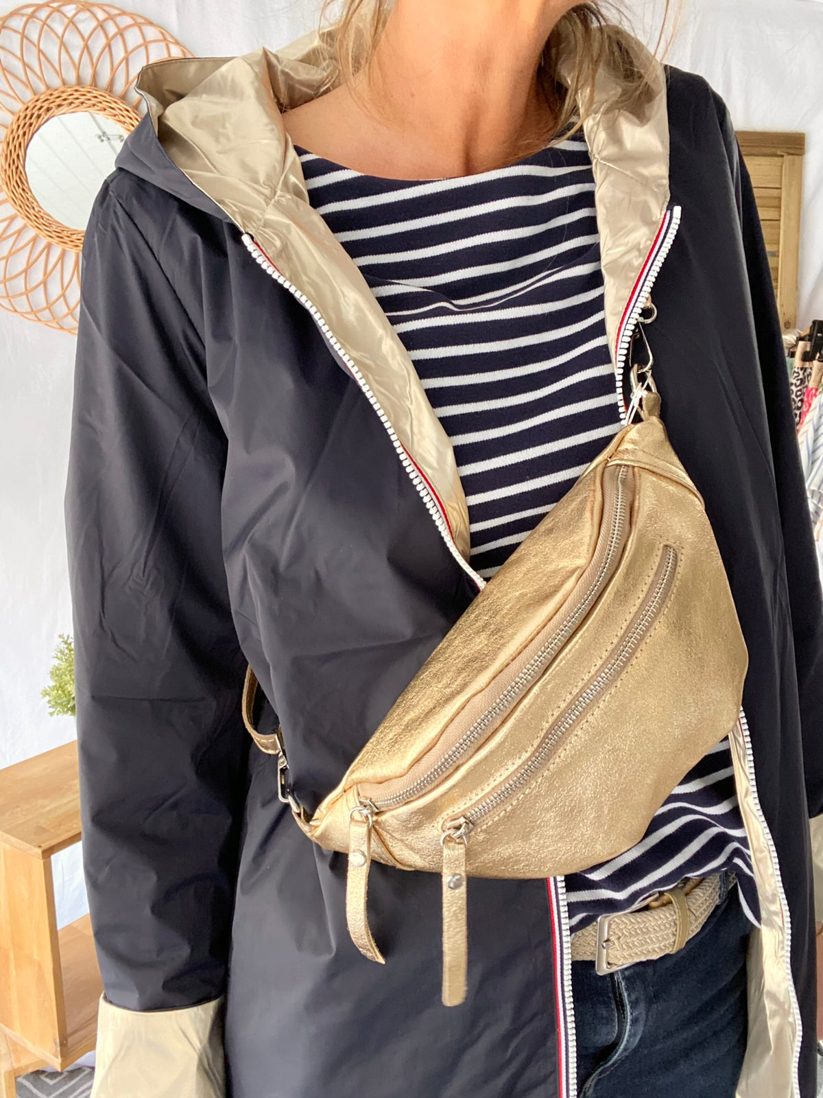 Femme portant un Sac banane en cuir irisé double poche - OPALE - Dorée avec sangle ajustable, sur une veste noire et chemise rayée.